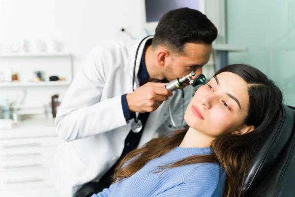 Sick young woman with an ear infection at the doctor\'s office sitting on an examination chair. Otolaryngologist doctor using an otoscope on a patient