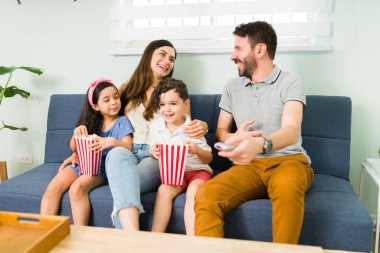 Çekici ebeveynler birbirlerine bakıp gülüyorlar. Küçük çocuklarıyla birlikte oturma odasında bir aile komedi filmi izliyorlar.
