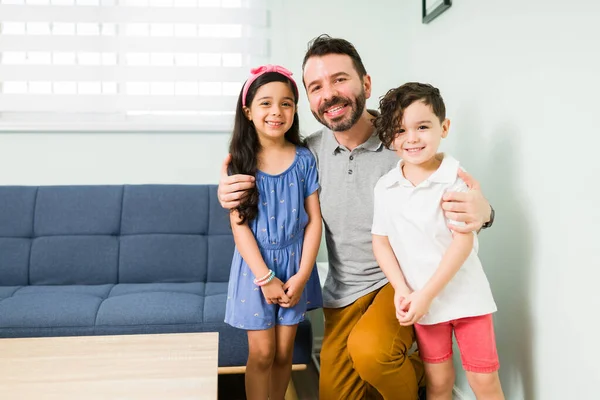 Portrait of a handsome dad hugging his little boy and girl while smiling while making eye contact at home. Latin father taking a picture with his children