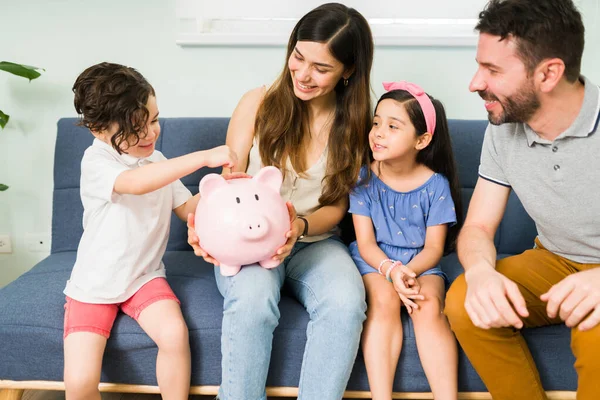 为家庭度假存钱 漂亮的四口之家坐在沙发上 把钱放在小猪存钱罐上 — 图库照片
