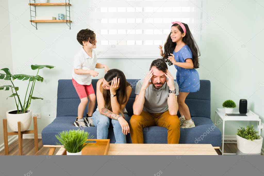 Stressed upset young parents feeling tired while their misbehaved children and jump on the couch around them 