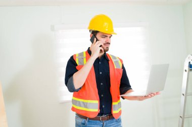 Portakal rengi anti-yansıtıcı yeleği olan odaklanmış mühendis dizüstü bilgisayardaki bir belgeyi okurken telefonla bilgi veriyor.