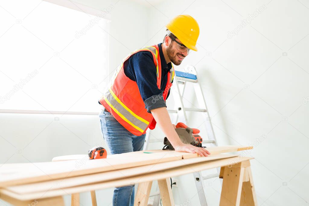 Happy male engineer with a safety helmet smiling while cutting a wooden panel with an electric machine