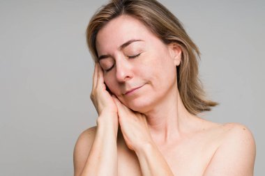 Rahat yaşlı bir kadın gözlerini kapatarak yüzüne dokunuyor. Beyaz kadın 30 'lu yaşlarda, gri bir stüdyo geçmişine karşı çıplak omuzlu.  