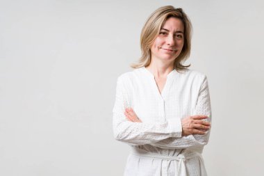 Gri stüdyo arka planında poz veren kolları katlanmış ve günlük kıyafetlerle gülümseyen muhteşem olgun bir kadın. 