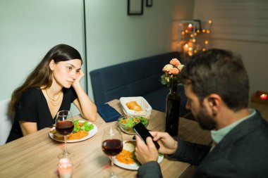 Canım çok sıkıldı. Sinirli genç kız arkadaşı, akşam yemeği sırasında akıllı telefonuyla mesajlaşan meşgul erkek arkadaşına kızgın bakıyor.