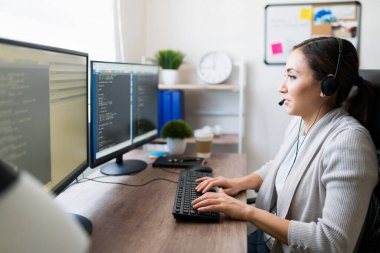 Evde ofis masasına yazılım kodu yazarken kulaklık takan ve bir müşteriyle konuşan mutlu bir kadın programcı.