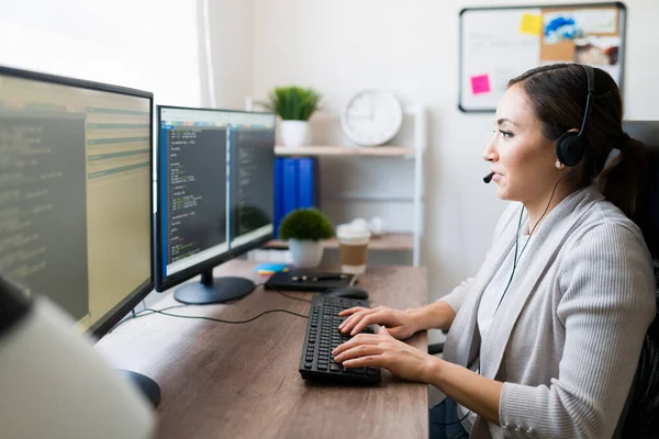 幸せな女性プログラマーはヘッドセットを着用し 自宅で彼女のオフィスの机の上にソフトウェアコードを書いている間 クライアントと話をする — ストック写真