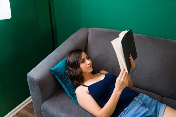 一个漂亮女人在沙发上看一本有趣的书的高度角度 三十多岁的女人带着一本小说躺在客厅里 — 图库照片