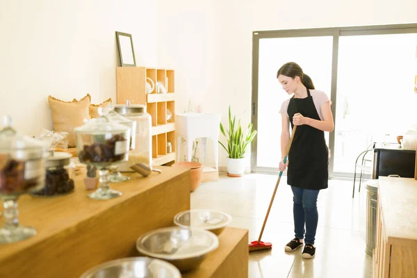 持続可能で廃棄されない食料品店の床を掃き掃除するエプロン付きの勤勉な女性労働者 — ストック写真