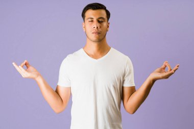 Sakin olun. Huzurlu genç bir adam, mor bir arka planda, gözleri kapalı meditasyon yapıyor ve rahatlıyor.
