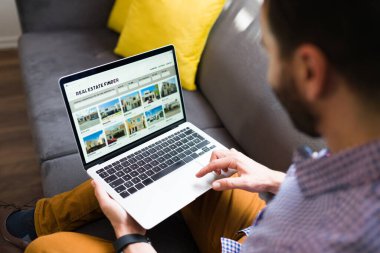 İnternette satılık yeni evler arayan bir adamın dikiz görüntüsü. Genç adam dizüstü bilgisayarında emlak sitesi kullanıyor. 