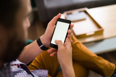 Genç insan evinde oturup boş bir ekrana bakarken akıllı telefonunu kullanıyor.