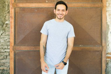 Latin genç adam gülümsüyor ve kameraya bakıyor. Gri bir tişört giyiyor.