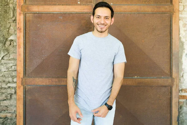 Latin genç adam gülümsüyor ve kameraya bakıyor. Gri bir tişört giyiyor.