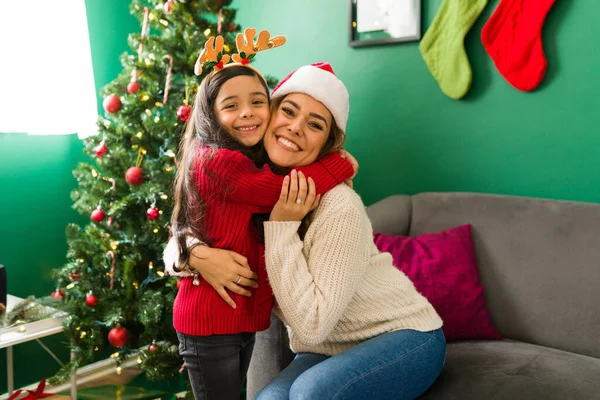 I love my mom. Hispanic little girl and cheerful young mom hugging while celebrating Christmas holiday at home