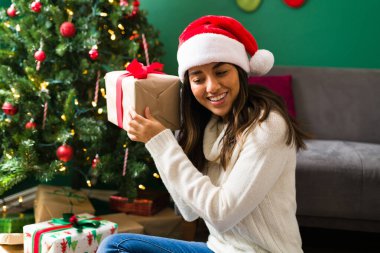 Noel Baba şapkalı süper mutlu kadın Noel hediyesini ağacın yanında tahmin etmeye çalışıyor.