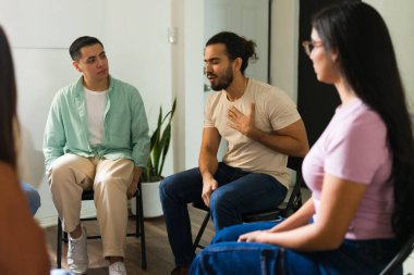 Genç adam, zihinsel sağlık destek grubu seansı sırasında bir çemberin içinde oturan, kişisel deneyimlerini dinleyen ve paylaşan insanlarla samimi bir hikaye paylaşıyor.