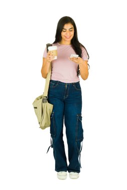 Latin bir kadın gülümsüyor ve yürüyor, elinde bir fincan kahve ve beyaz arka planda tam boy cep telefonu kullanıyor.