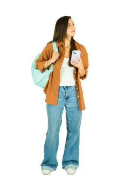 Sırt çantası ve akıllı telefonuyla duran genç kız öğrenci, beyaz stüdyo geçmişini araştırırken ya da planlarken düşünceli görünüyor.