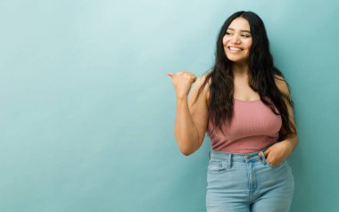 Pembe bluzlu ve kot pantolonlu neşeli İspanyol kadın arkasını gösteriyor. Terfi ya da reklam için boş stüdyo alanı gösteriyor.