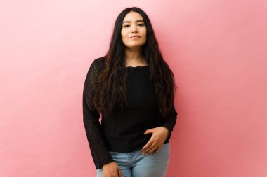 Uzun siyah saçlı İspanyol kadın kameraya bakıyor, stüdyoda duruyor, kendinden emin ve güçlü hissediyor.