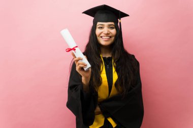 Şapka ve cübbe giymiş genç Latin mezunu. Pembe stüdyo geçmişine rağmen kameraya güvenle gülümsüyor.