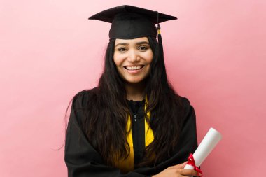 Eğitimini tamamladıktan sonra diplomasını elinde tutan gülümseyen genç bir kadın. Stüdyoda mezuniyet kepi ve cüppe takıyor.
