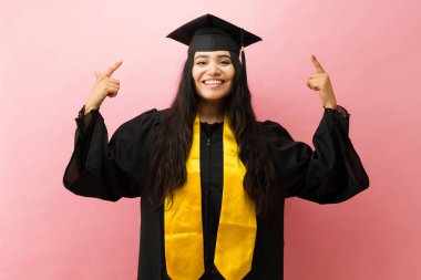 Genç kız öğrenci gülümsüyor, mezuniyet elbisesi giyiyor, şapka takıyor ve fotokopi alanını gösterirken çalıyordu.