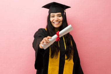 Mezuniyet cüppeli mutlu Latin Amerikalı bayan öğrenci pembe bir stüdyo geçmişine karşı akademik diploma taşıyor.