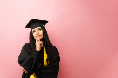 Genç bir bayan öğrenci kep ve cübbe giyiyor, mesleki yolunu düşünürken başını kaldırıyor.