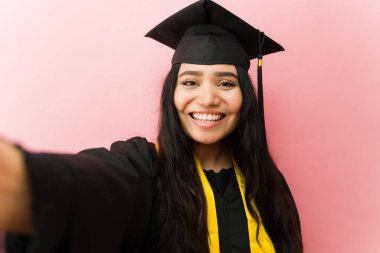 Genç bayan mezun, başarıyı kutluyor. Pembe arka planda kep ve cübbe giyerek selfie çekiyor.