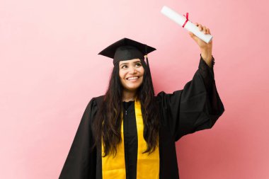 Akademik giyinmiş, şapkalı gülümseyen genç bir kadın, diploma yetiştirmek, mezuniyeti pembe bir arka planda kutlamak.