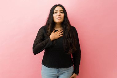 Hispanik kadın göğsünde ağrı ya da rahatsızlık hissediyor, elini kalbinin üstünde tutuyor, endişeli ve pembe arka planda endişeli görünüyor.