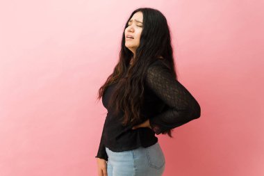 Sırt ağrısı çeken Latin bir kadın stüdyoda acı dolu bir ifadeyle sırtını tutuyor.