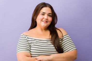 Kollarını kavuşturup kameraya gülümseyen iri yarı bir kadın mor stüdyo arka planında vücut pozitifliğini kutluyor.