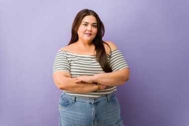 Çizgili omuzlu, kot pantolonlu, kollarını kavuşturmuş gülümseyen kıvrımlı kadın vücut pozitifliğini ve kendine güvenini kutlayan stüdyo portresi.