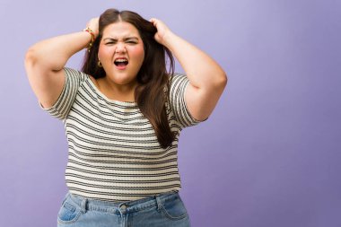 Genç ve cüsseli bir kadın saçını çekiyor, bağırıyor, hayal kırıklığı, öfke ve stres gösteriyor, stüdyoda duruyor.