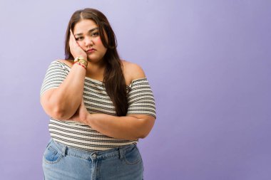 Stüdyoda kendini sıkılmış ve üzgün hisseden genç ve cüsseli bir kadın melankolik bir ifadeyle elini yüzüne koyuyor.