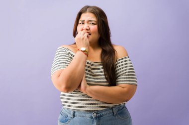 Büyük beden bir kadın endişeli ve endişeli hissediyor, tırnaklarını yiyor. Mor bir arka planda korku ve gerginlik gösteriyor.