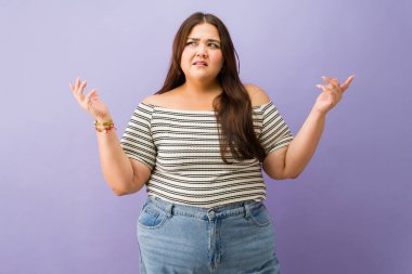 Kıvrımlı Latin kadın, kafası karışık ve omuzlarını silkiyor. Mor stüdyo geçmişini bilmemenin jestini yapıyor.