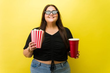 Üç boyutlu gözlüklü, elinde patlamış mısır ve soda ile gülümseyen, sarı arka plana karşı eğlenceli bir sinema anının tadını çıkaran iri yarı bir kadın.