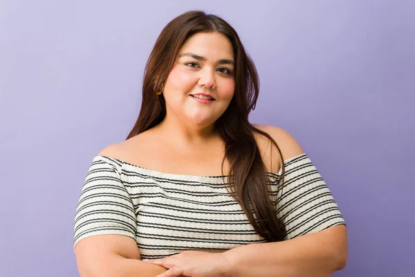 Kollarını kavuşturup kameraya gülümseyen iri yarı bir kadın mor stüdyo arka planında vücut pozitifliğini kutluyor.