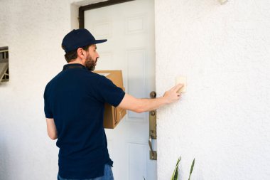 Üniformalı ve şapkalı bir erkek kurye karton bir paket getiriyor, müşteri sevkiyatı için ön kapı zilini çalıyor.