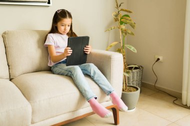 Evdeki kanepede dinlenirken dijital tablete konsantre olan genç kız, e-öğrenme ve ekran zamanının keyfini çıkarıyor.