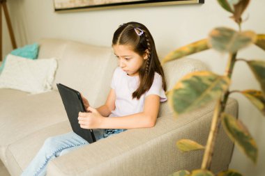 Kanepede oturan genç bir kız, eğitim ve eğlence için tabletle meşgul, kapalı alanda eğleniyor.