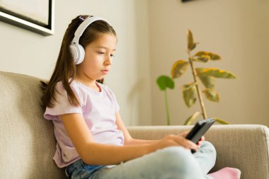 Küçük çocuk kanepede oturuyor, akıllı telefon ekranına konsantre olurken kablosuz kulaklıkla ses dinliyor, teknolojiyle bağlantı kuruyor.