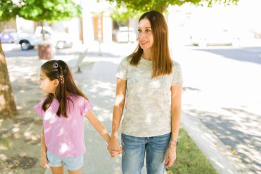 Anne ve kızı kaldırımda yürürken el ele tutuşuyorlar, güneşli bir günde dışarıda güzel vakit geçiriyorlar.