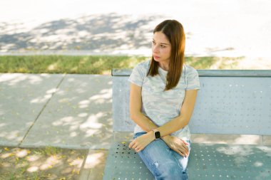 Parktaki bankta tek başına oturan bir kadın, dalgın bir ifadeyle başka yere bakıyor, üzgün ve yalnız hissediyor.