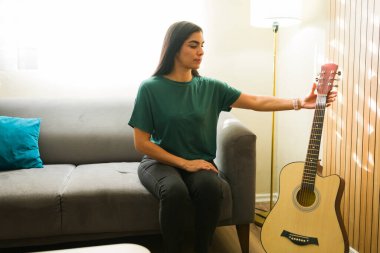 Kanepede oturan bir kadın, oturma odasında akustik gitar çalmaya hazırlanıyor, müzik hobisinin tadını çıkarıyor.